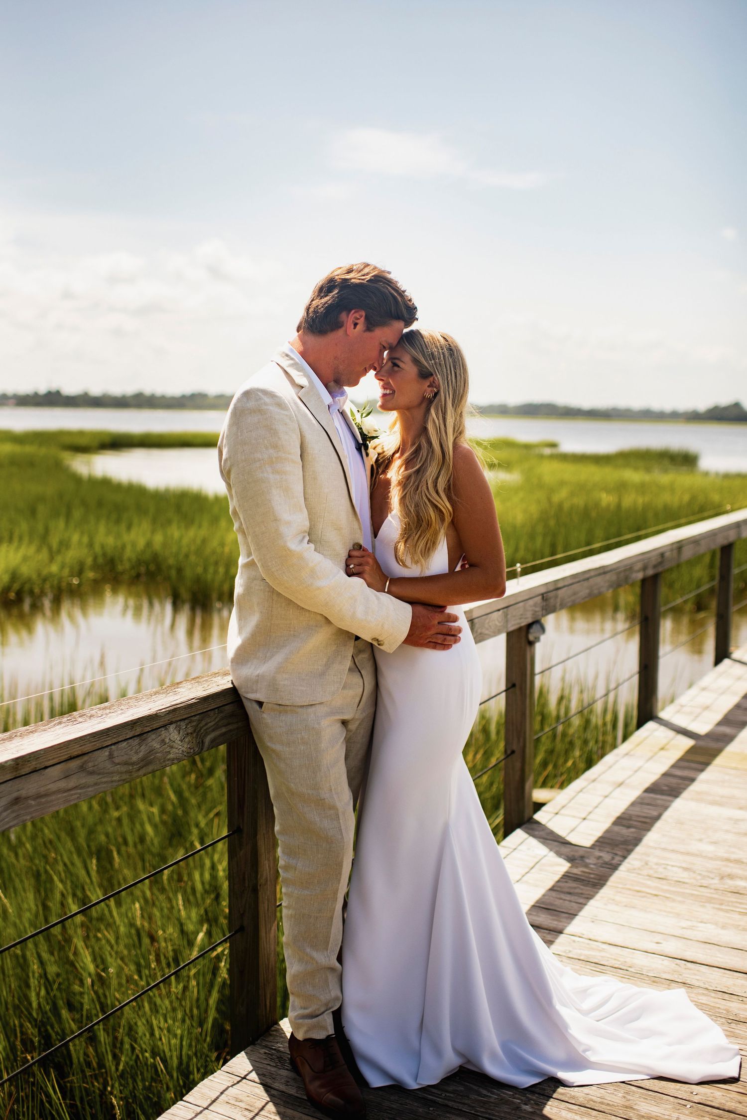 A romantic wedding couple embraces on a wooden boardwalk overlooking coastal marshlands at sunset.