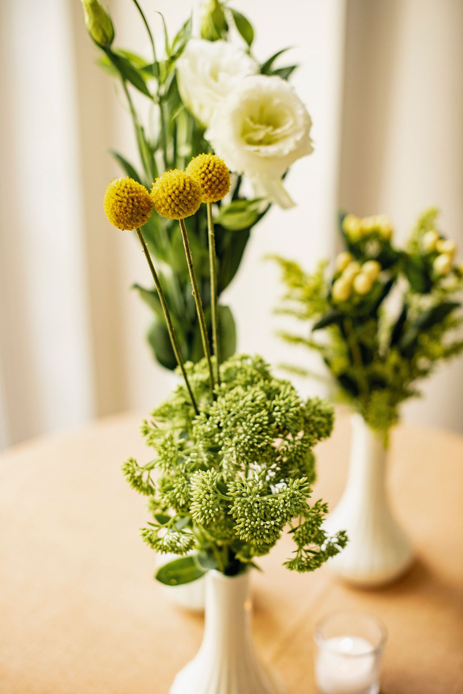 White flowers and yellow craspedia arranged with delicate green foliage in a minimalist white ceramic vase.