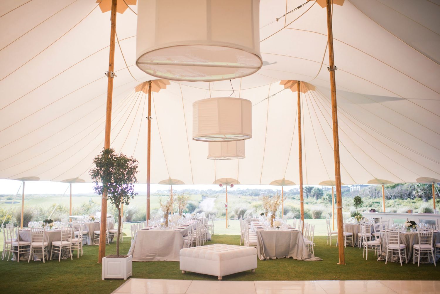 Elegant white tent wedding reception setup with hanging drum shade chandeliers and round tables on a grassy lawn.