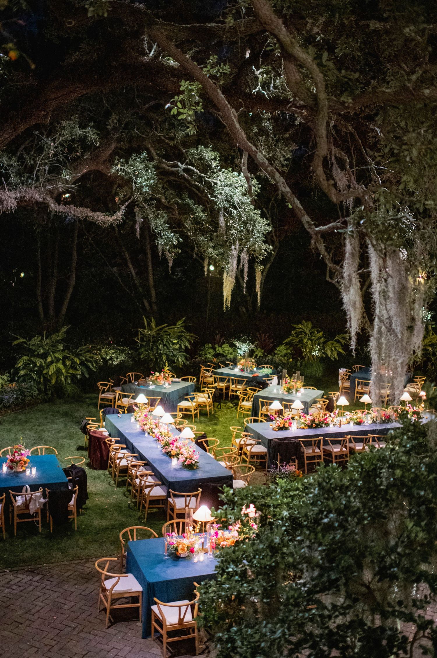 Outdoor evening wedding reception with illuminated long tables set under Spanish moss-draped oak trees in a garden setting.