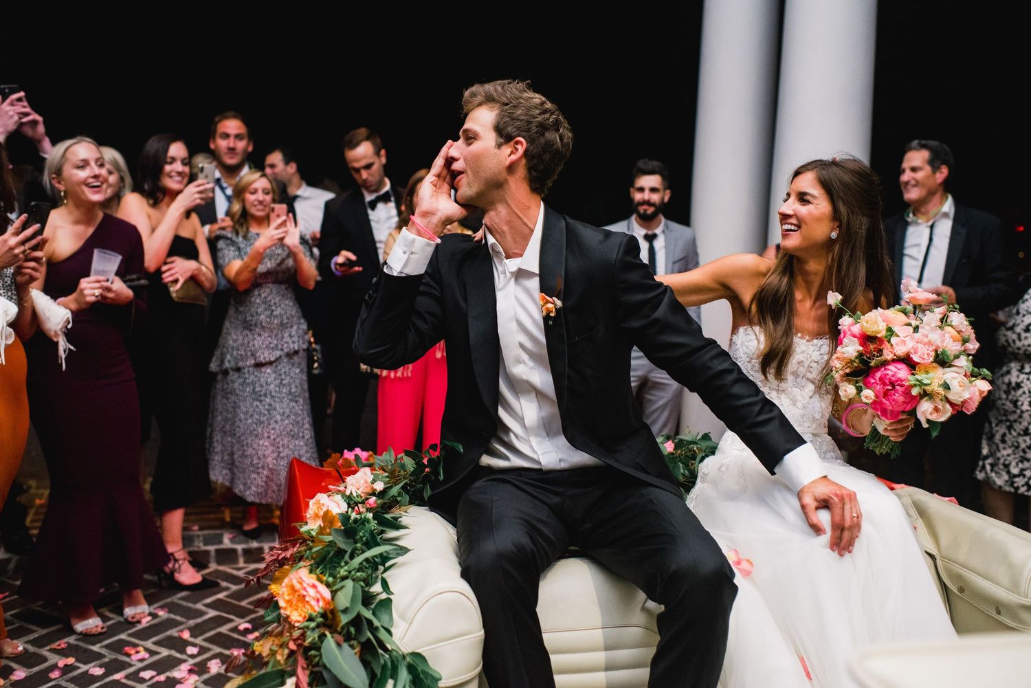 Wedding guests celebrate and dance during a lively reception at an elegant venue with white columns.