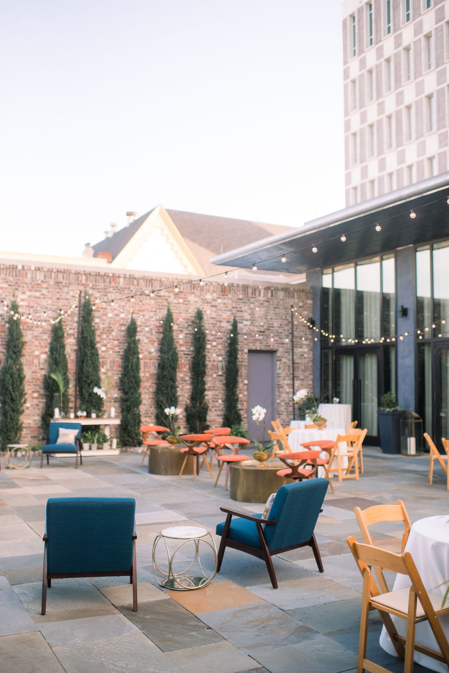 Modern outdoor patio space with blue lounge chairs, wooden furniture, and string lights against a stone wall backdrop.