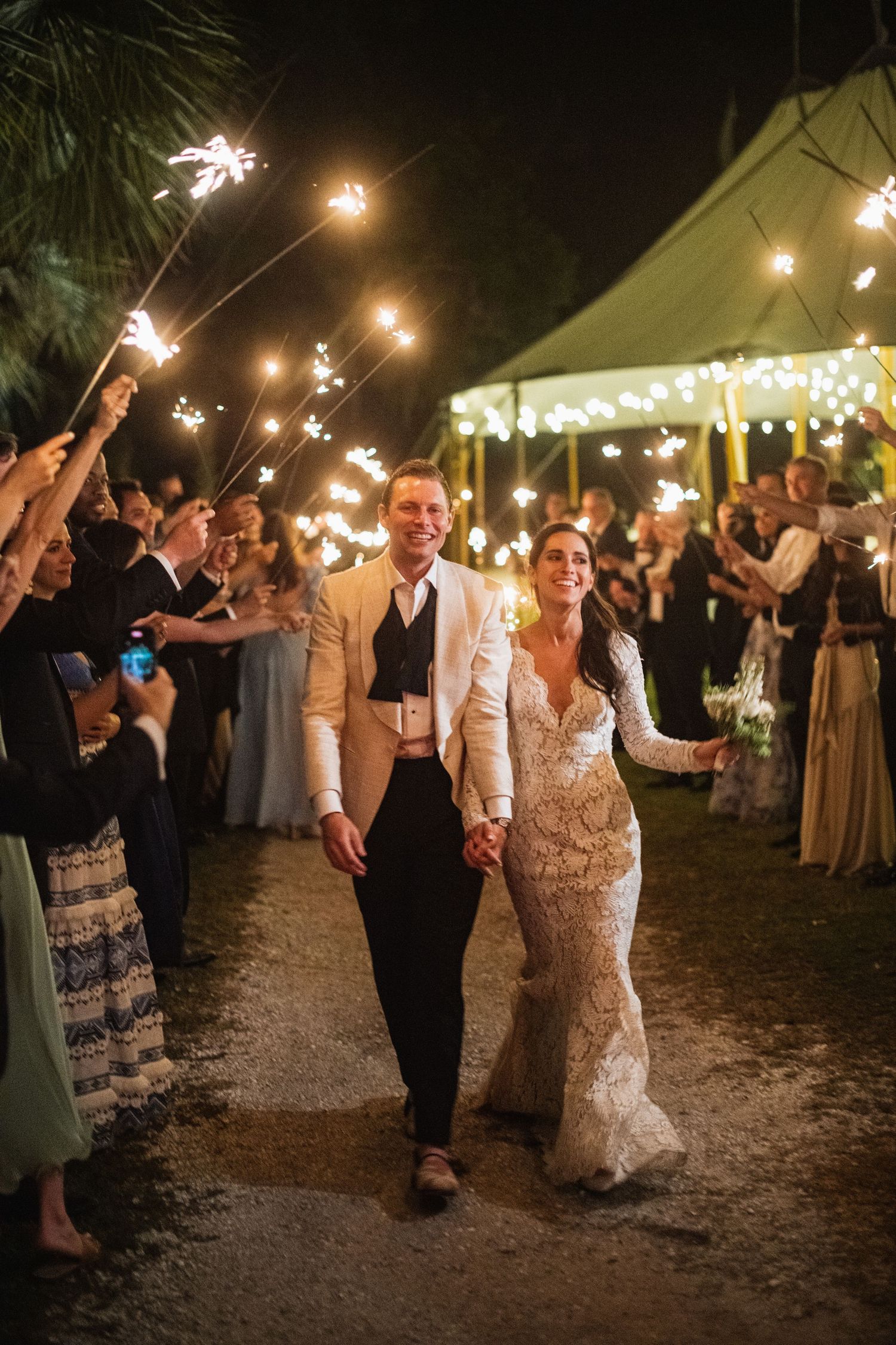 Wedding guests celebrate under string lights at an outdoor evening reception as couple dances through sparkler exit.