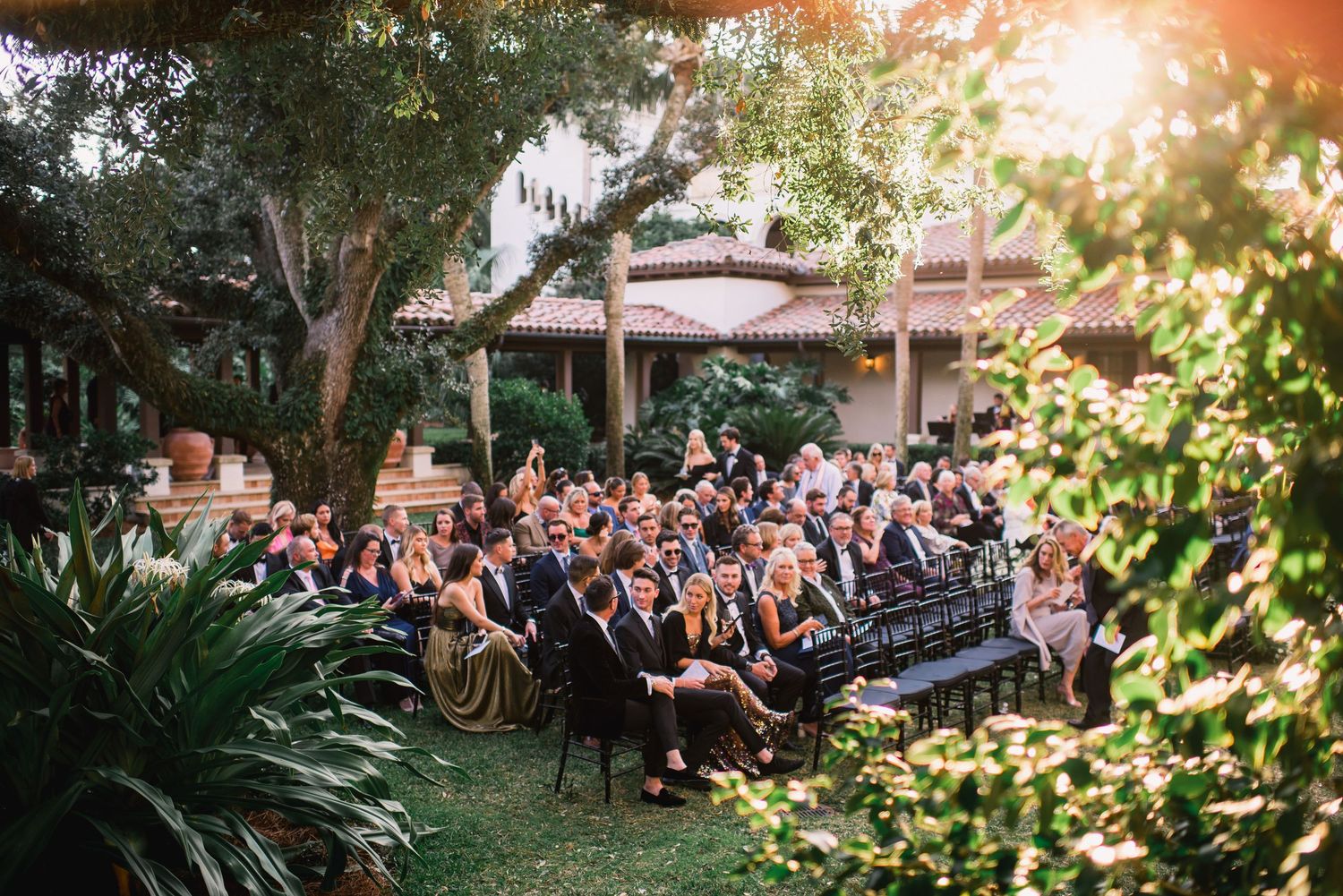Guests gather outdoors at a sunset wedding reception with string lights illuminating the garden space.