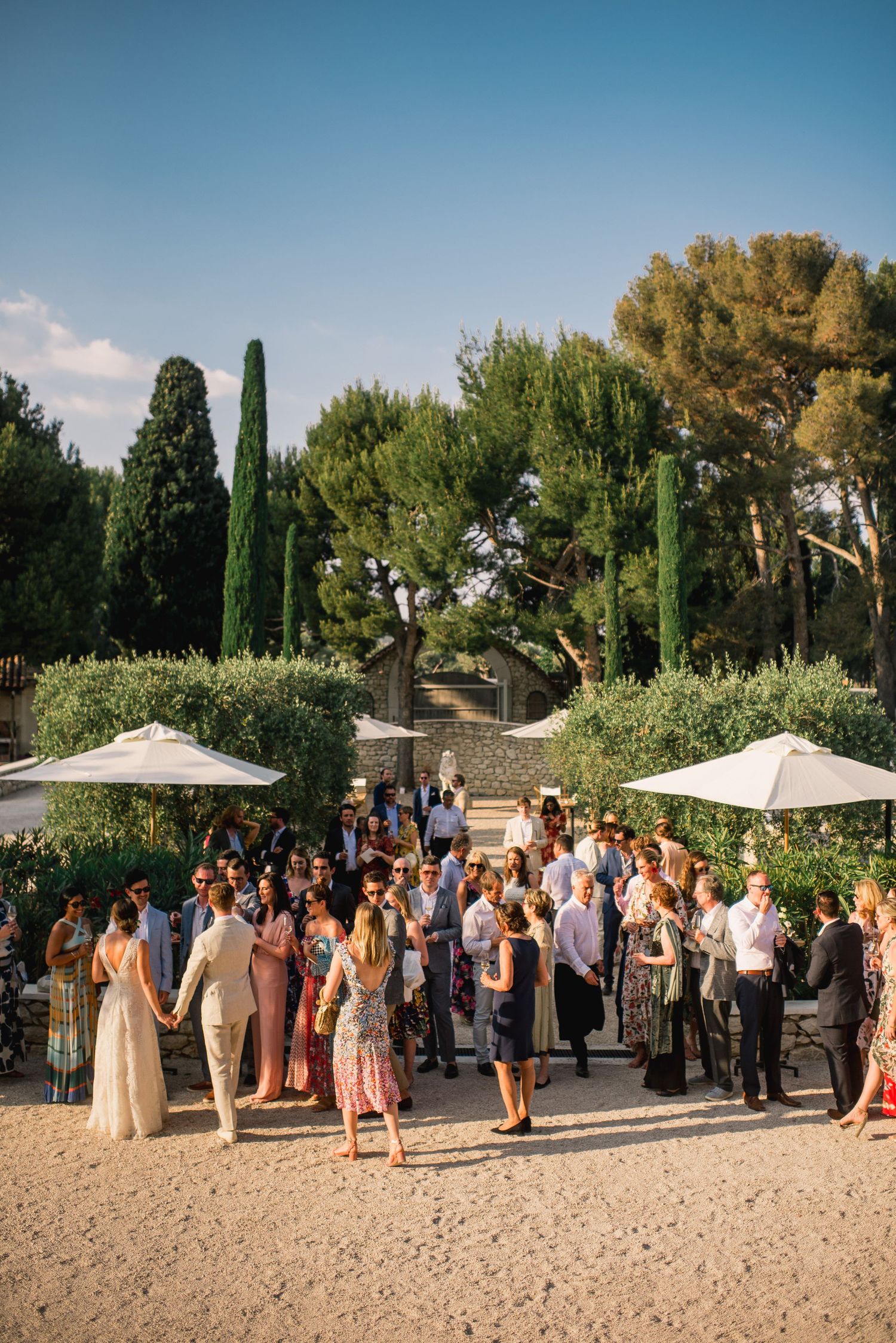 A large group gathers for an outdoor event in a Mediterranean garden setting with cypress trees and white umbrellas.