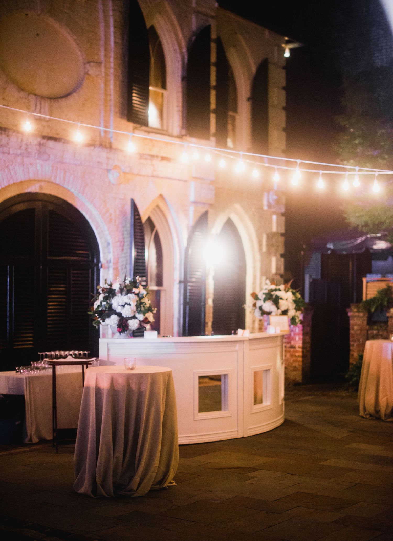 String lights illuminate an elegant outdoor evening reception area with white-draped tables and classic stone architecture.