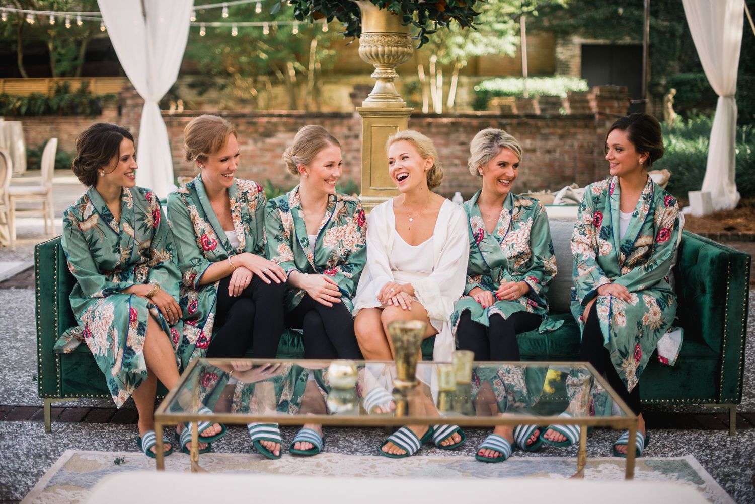 Wedding party in turquoise dresses and suits laughs together while sitting on stone wall at outdoor reception.