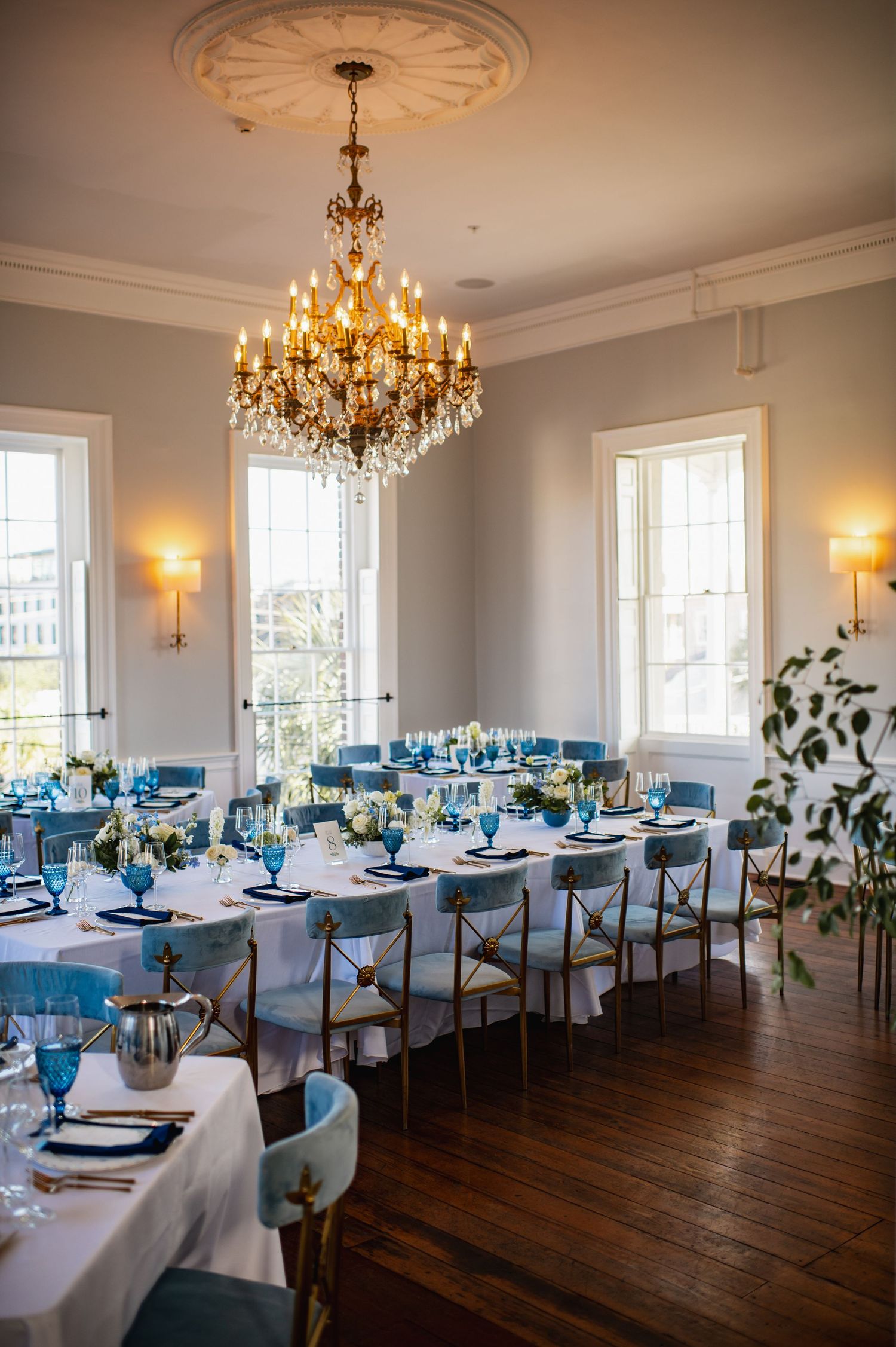 Elegant dining room setup with blue and white table settings, crystal chandeliers, and tall windows in a historic venue.