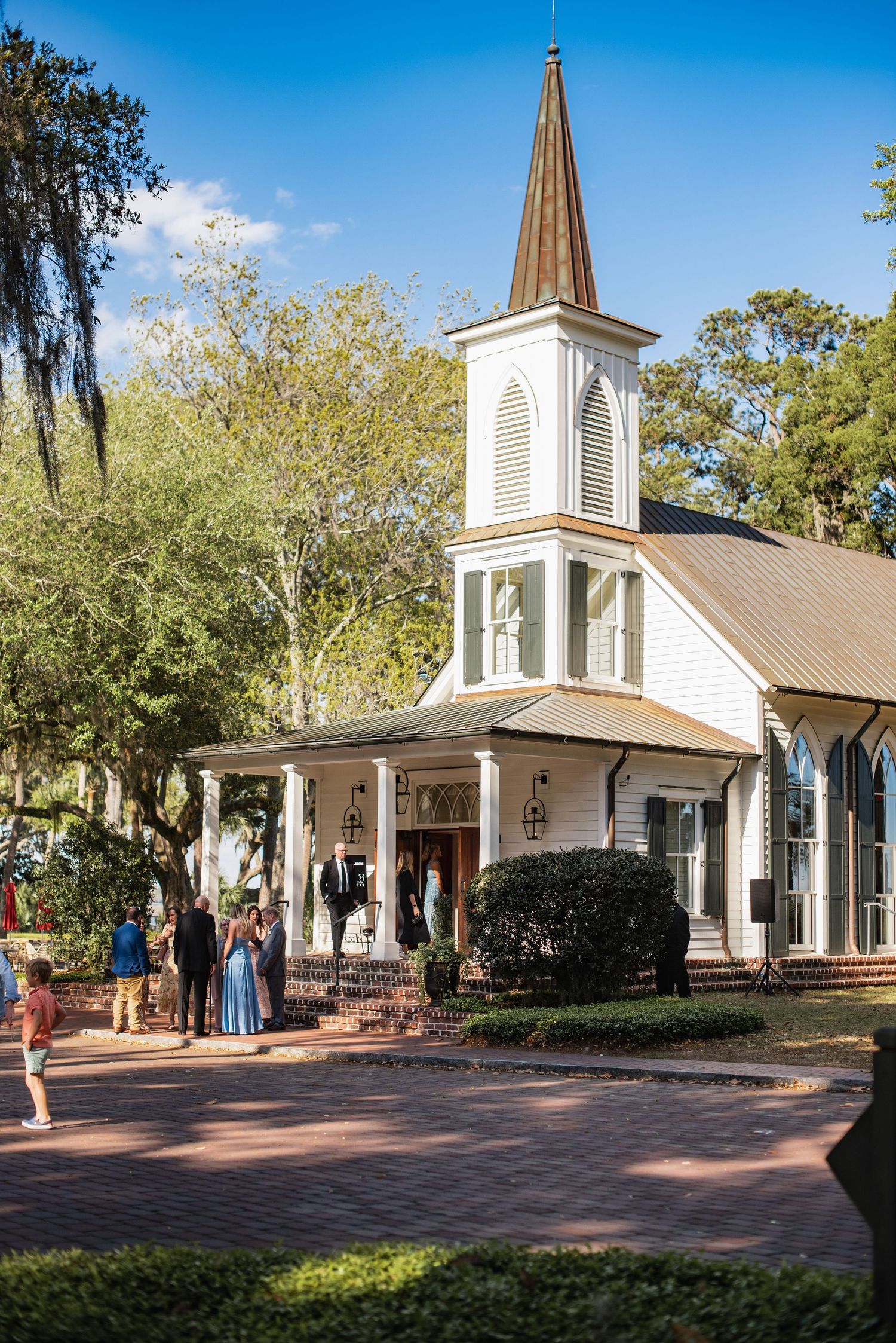 A charming white chapel with a tall steeple stands among Spanish moss-draped trees on a brick pathway.