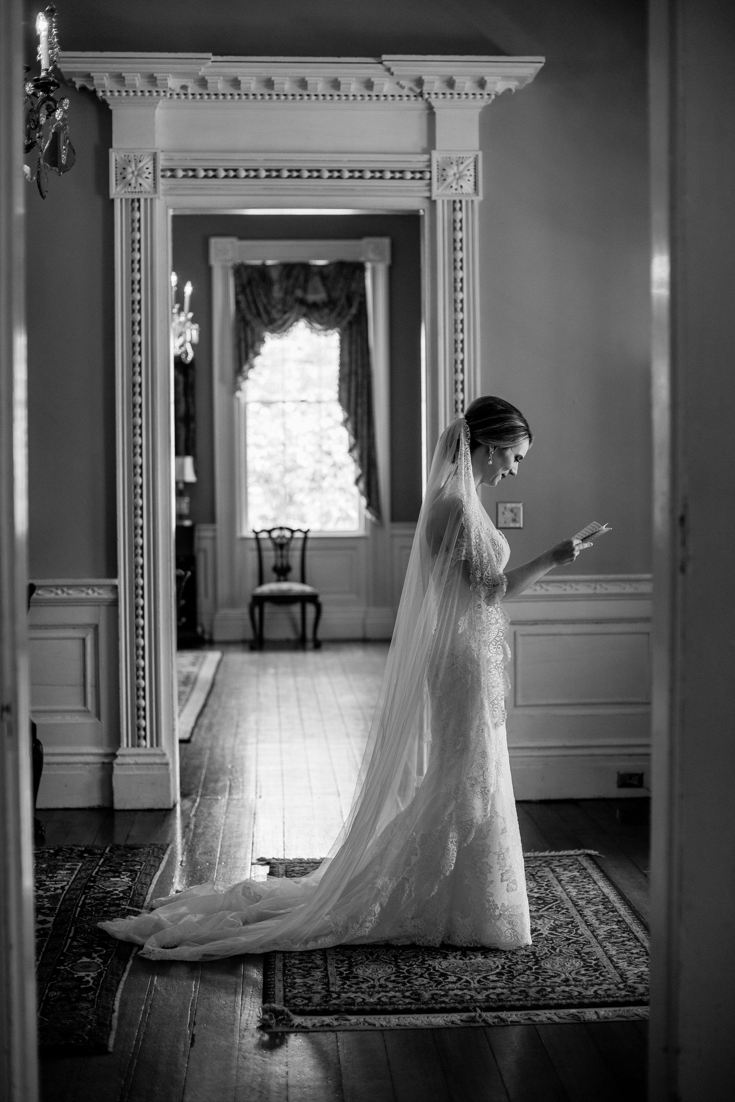A bride in a long lace dress and veil stands in an elegant room with ornate molding and a window with flowing curtains.