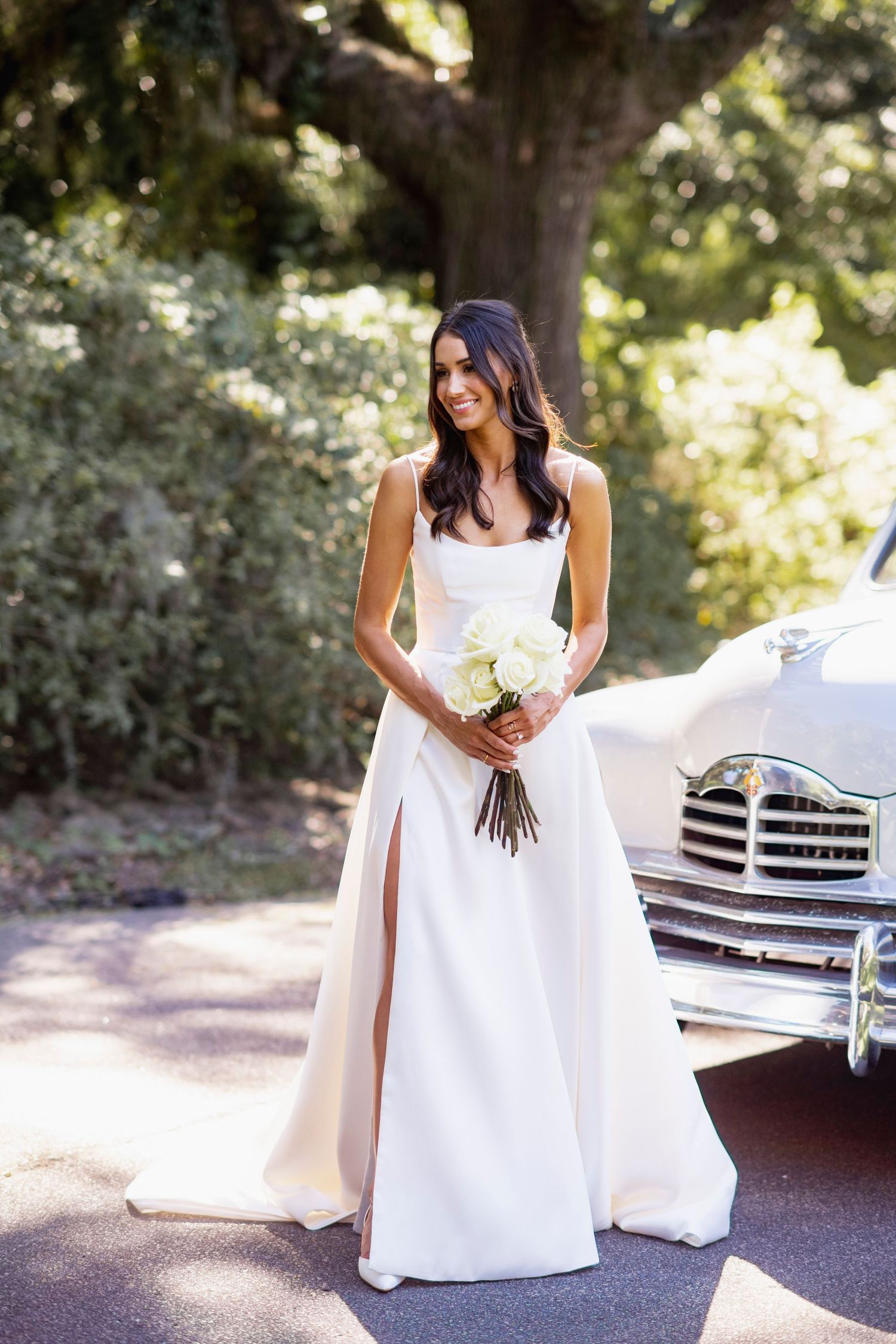Elegant bride in white wedding dress poses gracefully in a sunlit outdoor setting with classic benches.