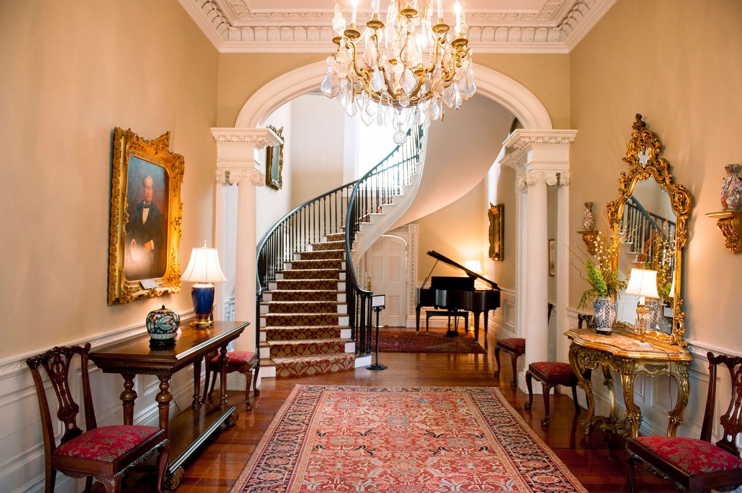 Elegant mansion interior with crystal chandelier, ornate moldings, and grand piano in a luxurious hallway with red carpet.