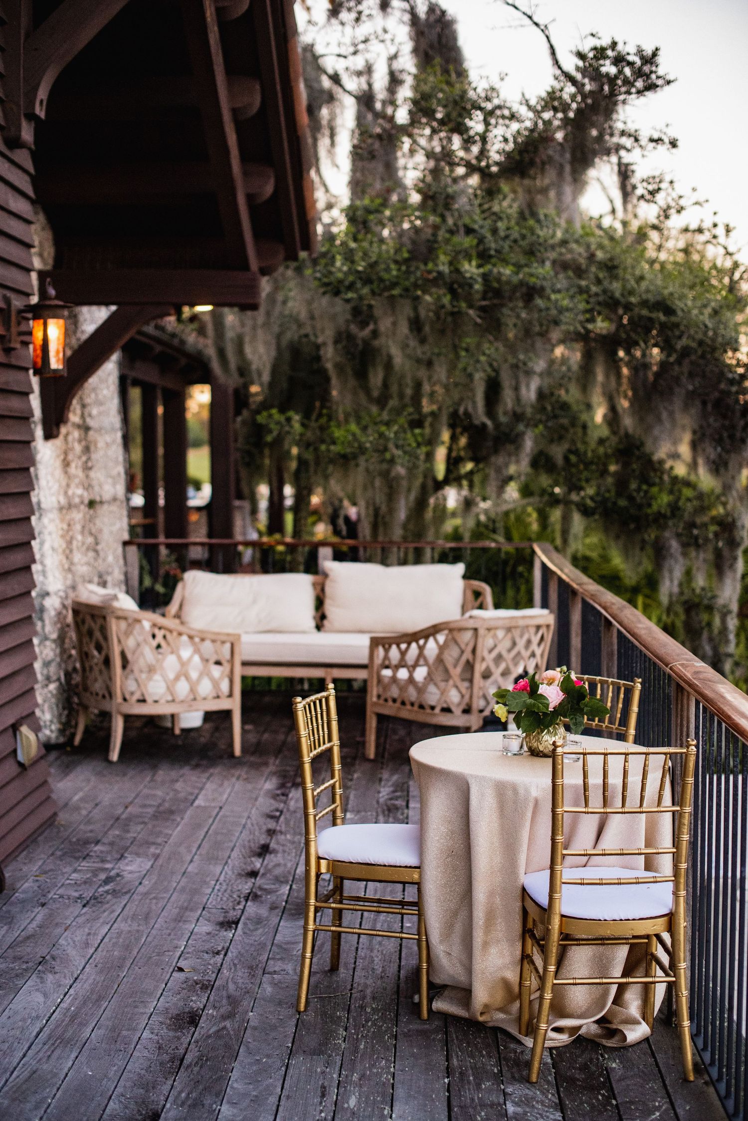 Rustic outdoor porch with vintage bamboo chairs and white cushions arranged against wooden railings and Spanish moss backdrop.