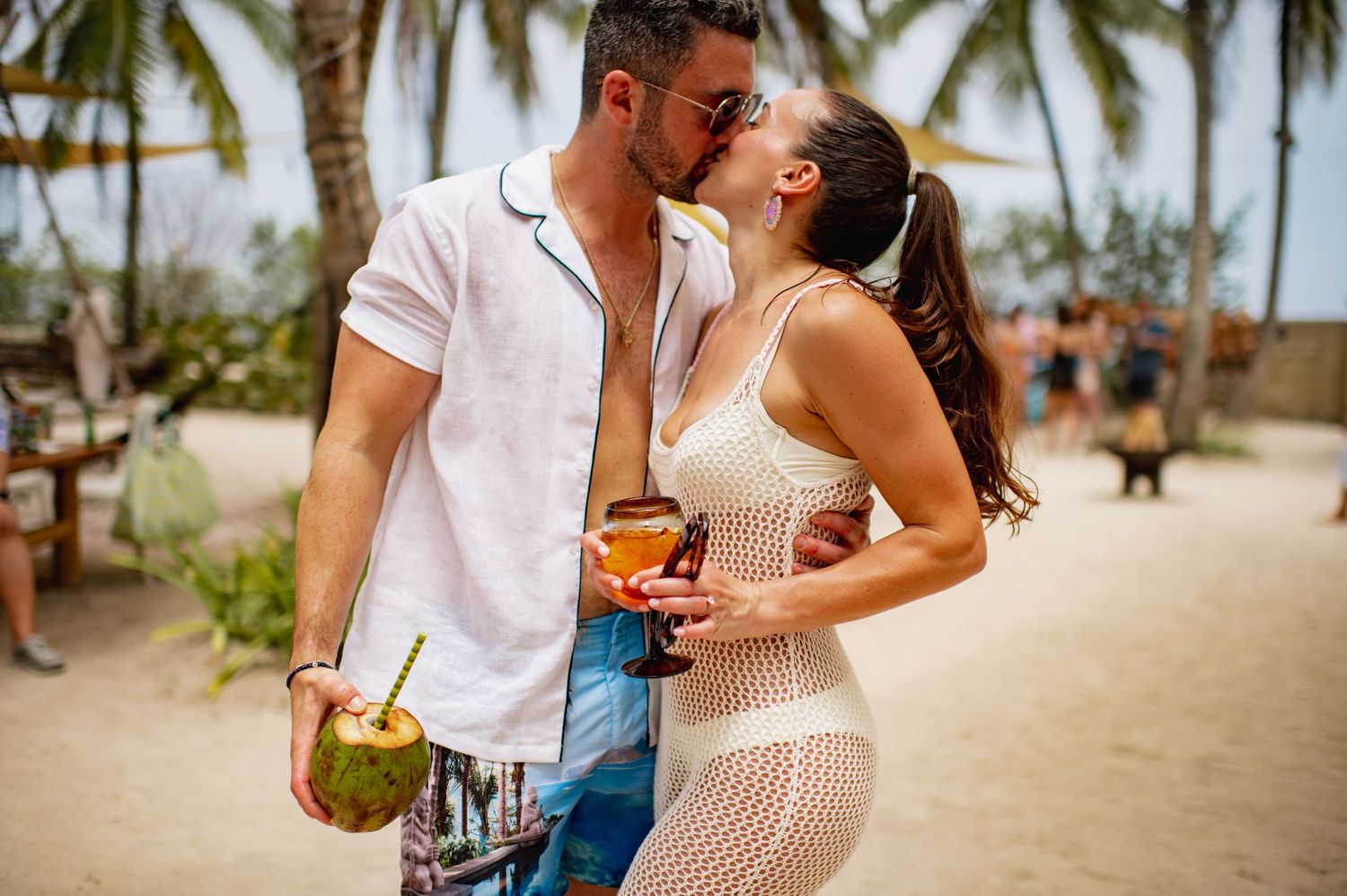 Couple in casual attire sharing romantic moments on a tropical beach with palm trees in the background.