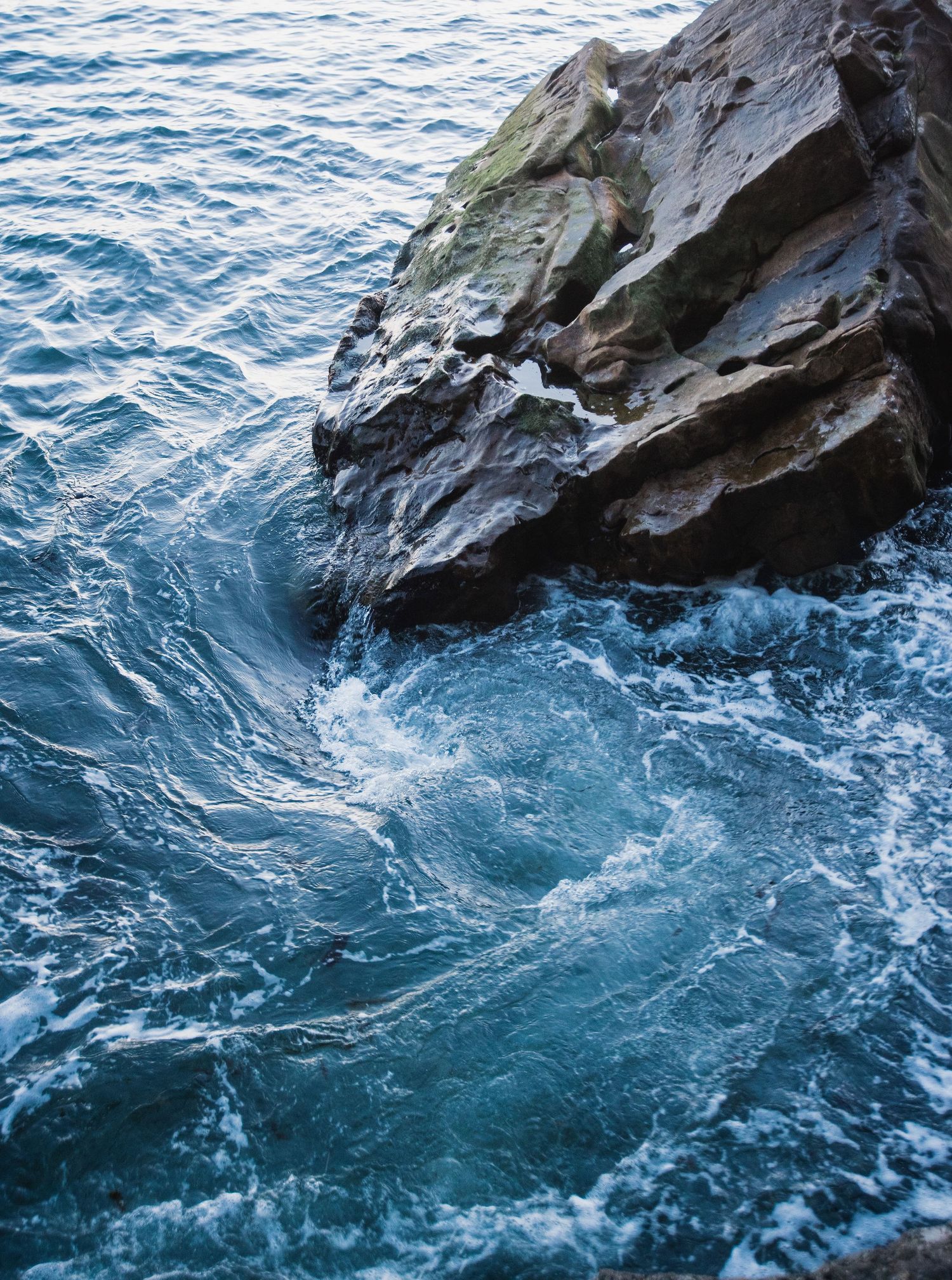 Waves crash against a rugged rocky cliff face creating swirling patterns in the deep blue ocean water.
