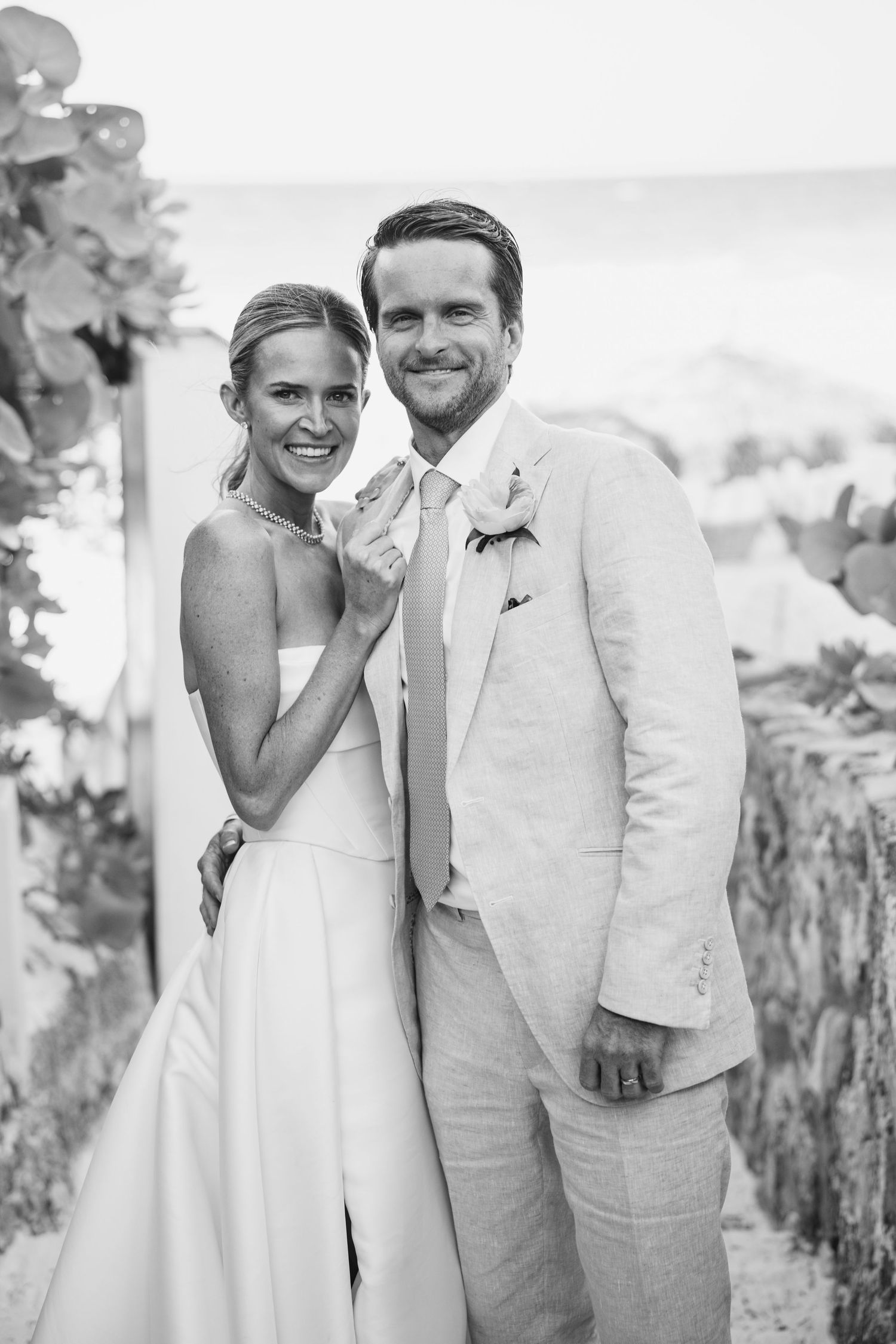Black and white portrait of newlyweds in formal attire against scenic coastal backdrop.