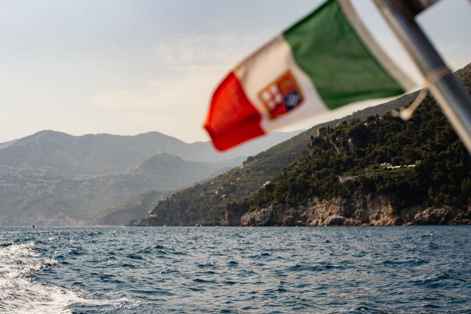 Italian flag waves over blue waters with in Amalfi coast in the background.