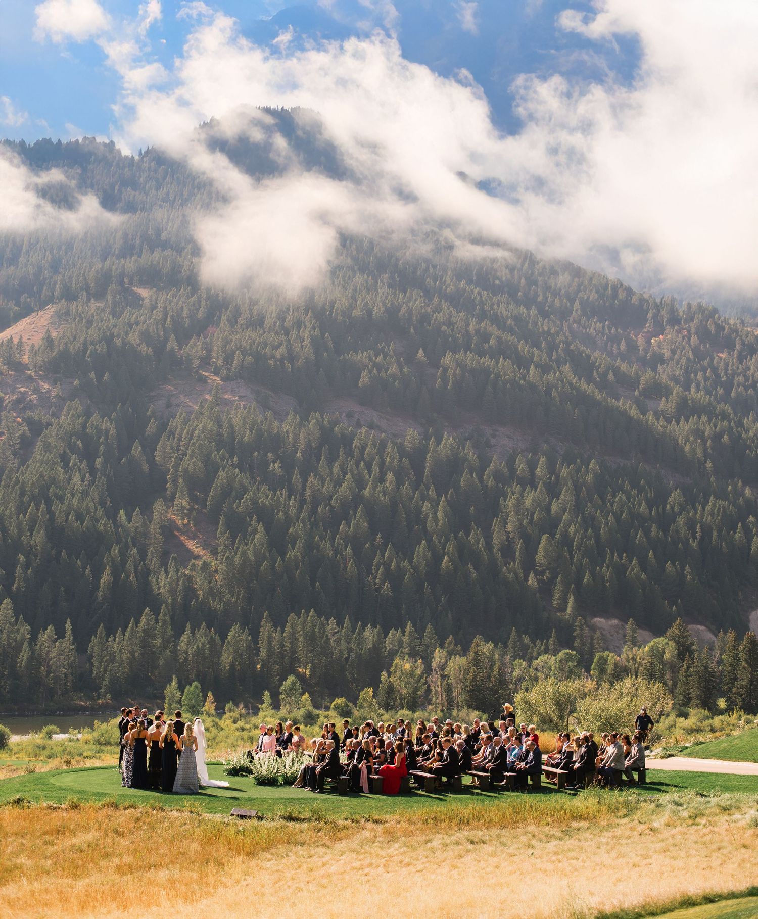 A large outdoor gathering in a meadow surrounded by misty mountain slopes covered in evergreen forest.