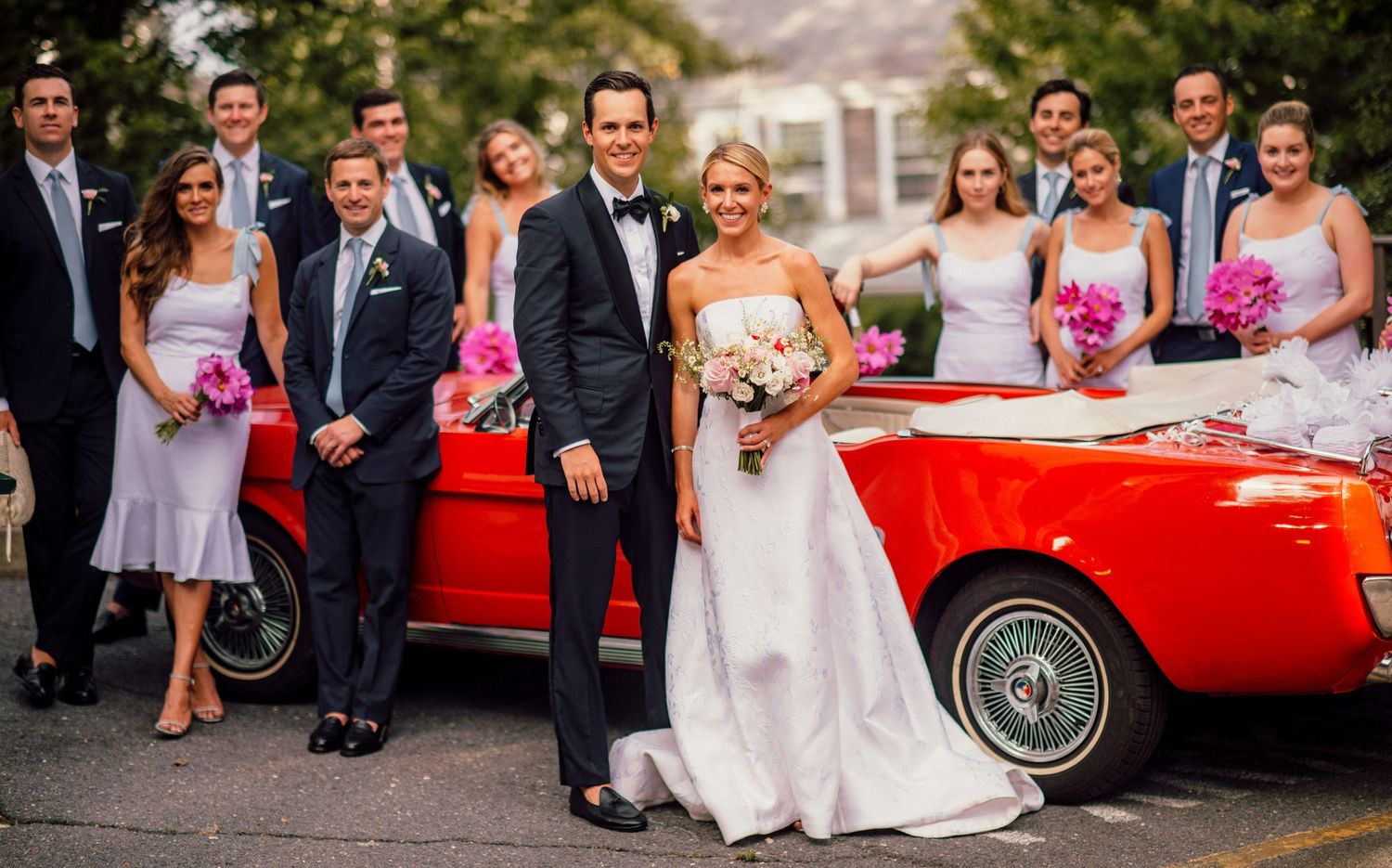 Wedding party poses next to a classic red convertible car outdoors.