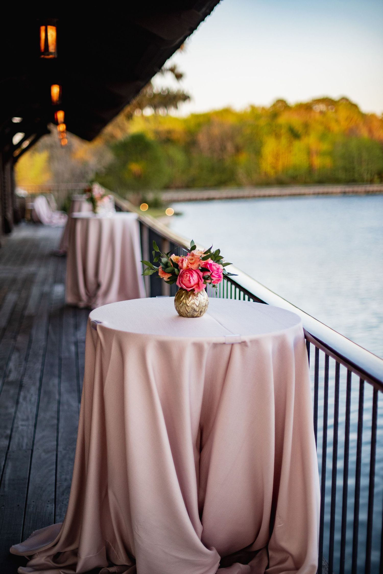 Elegant cocktail tables with blush pink linens and floral centerpieces set up on a waterfront deck at sunset.