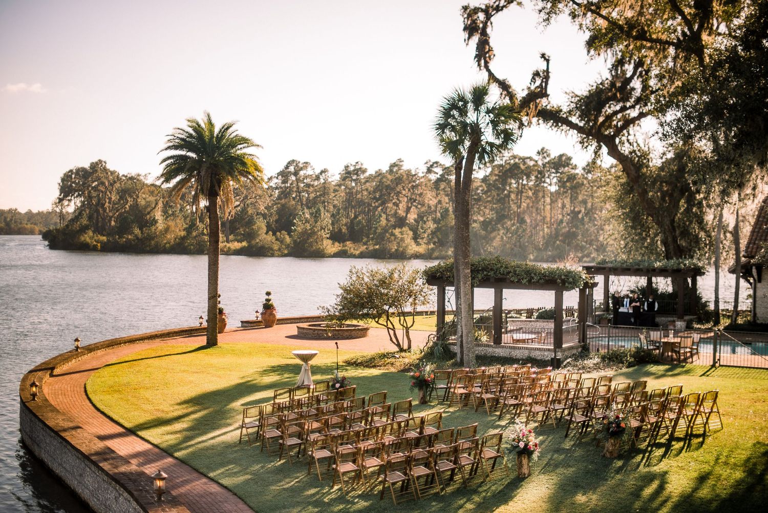 Outdoor wedding ceremony setup with rows of chairs on a waterfront lawn surrounded by palm trees at dusk.