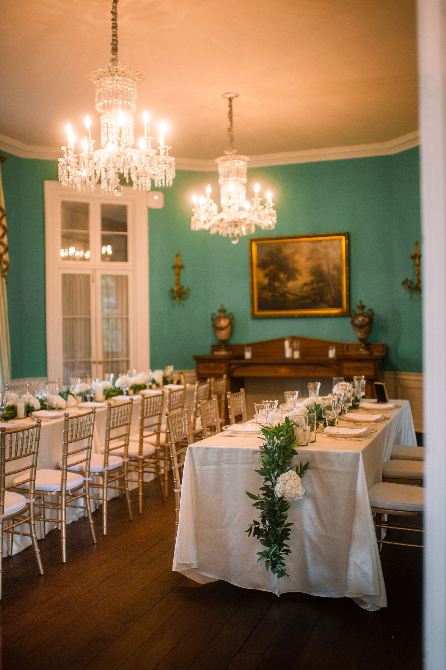 Elegant dining room with turquoise walls, crystal chandeliers, and long tables set with white linens for a formal event.