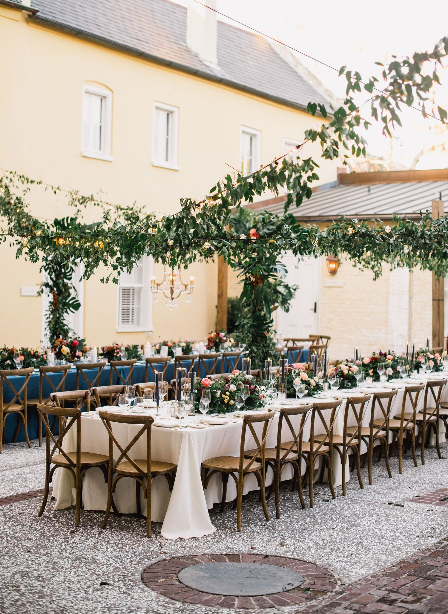 Elegant outdoor wedding reception setup with wooden cross-back chairs and white tablecloths under string lights and greenery.