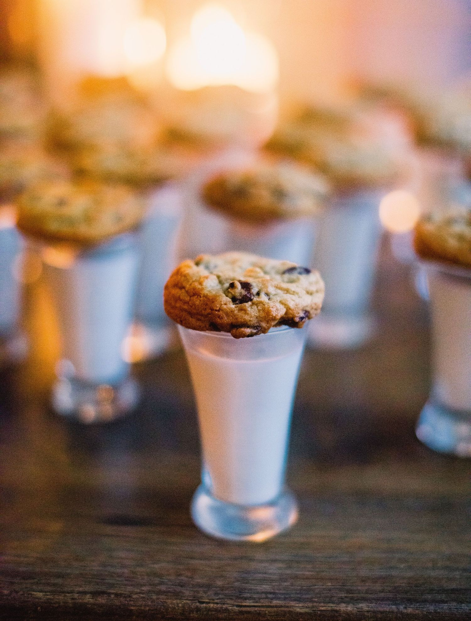 Chocolate chip cookie balanced on the rim of a small white milk cup with blurred cookies in background.