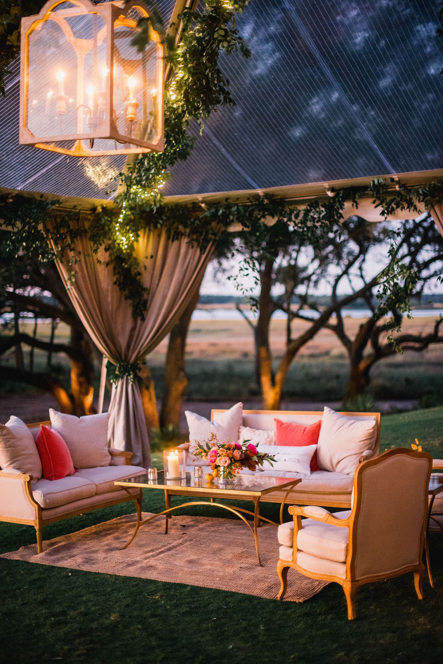 Elegant outdoor lounge area with white cushioned seating, draped curtains, and hanging lanterns at sunset.