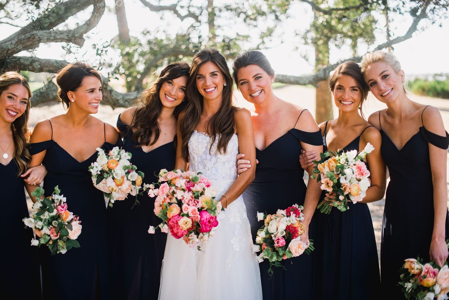 Wedding party members in black dresses hold colorful bouquets while posing outdoors for group photos.