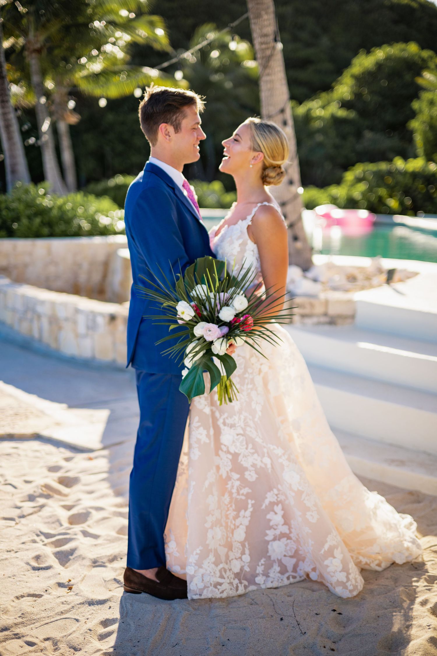 Newlyweds pose together on a sandy beach with tropical flowers and palm trees.