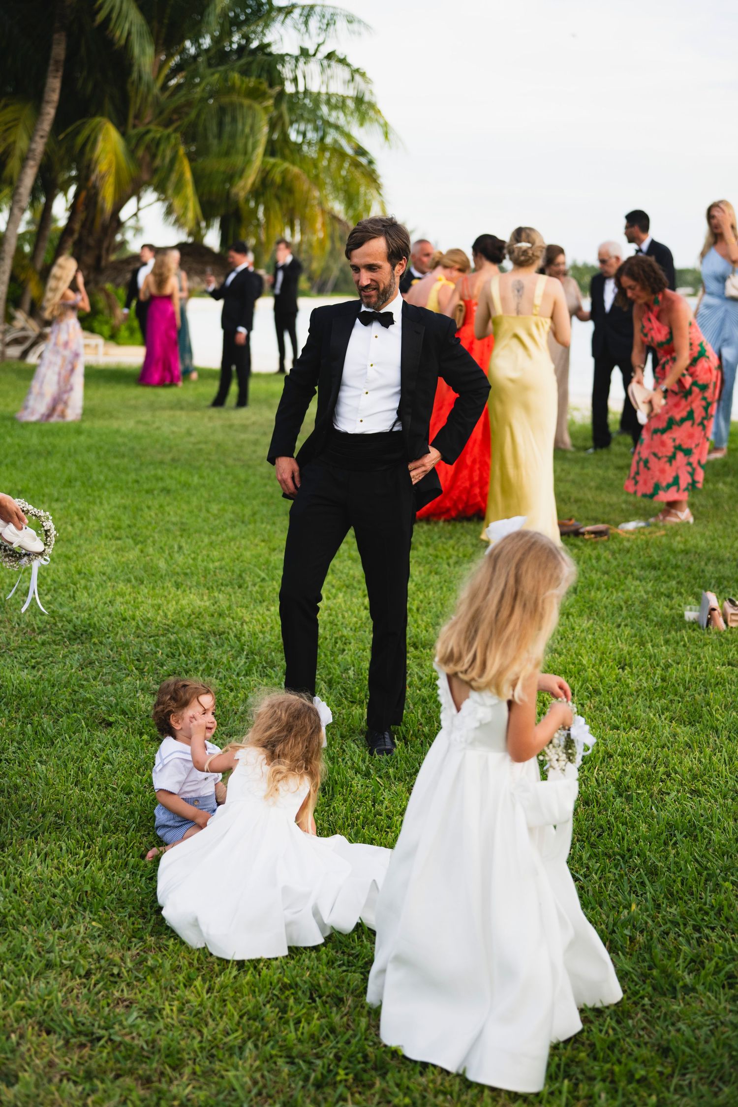 Children in formal attire play and run on a grassy lawn during an outdoor wedding reception.