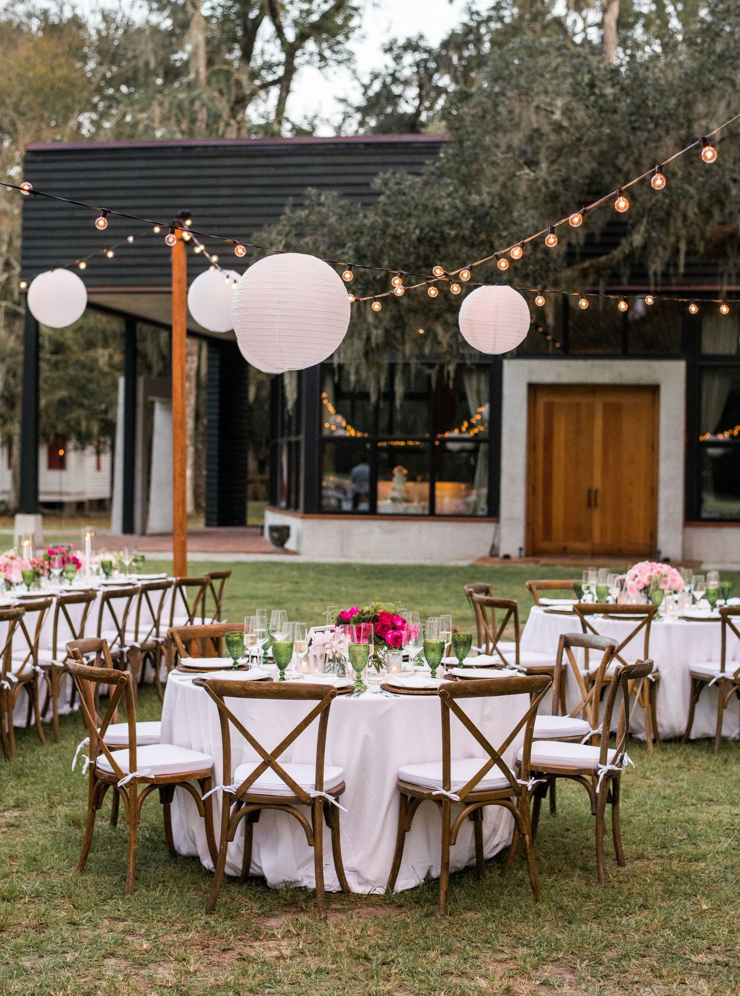 Elegant outdoor wedding reception setup with round tables, wooden chairs, white linens, and hanging paper lanterns and string lights.