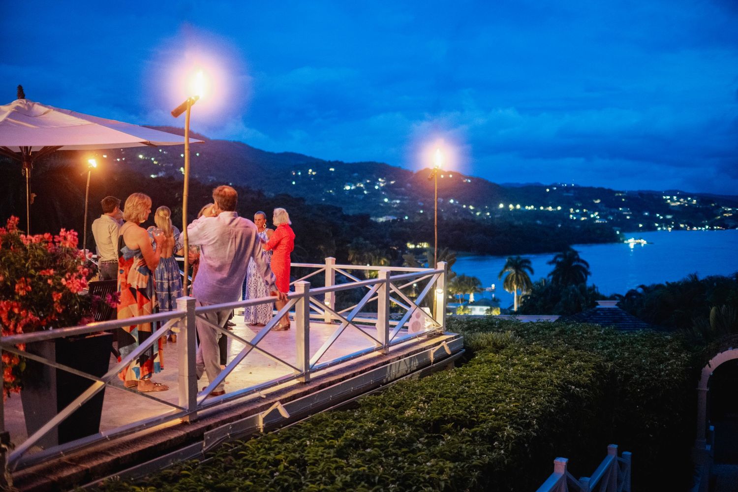 Evening wedding guests gather on an illuminated deck overlooking a scenic waterfront bay at dusk.