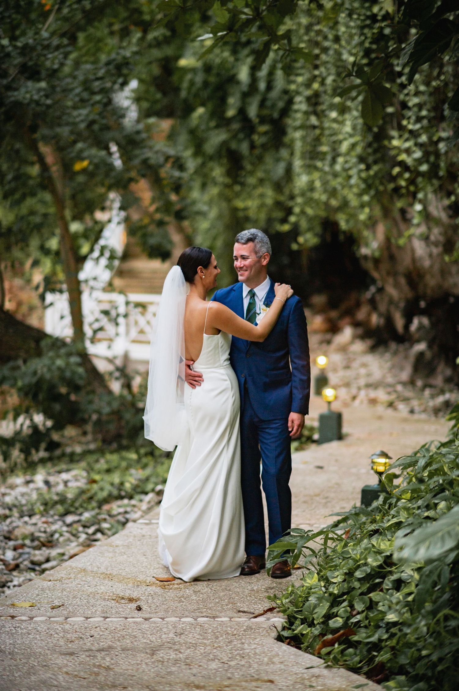 A couple in wedding attire sits on stone steps surrounded by lush greenery in an intimate garden setting.
