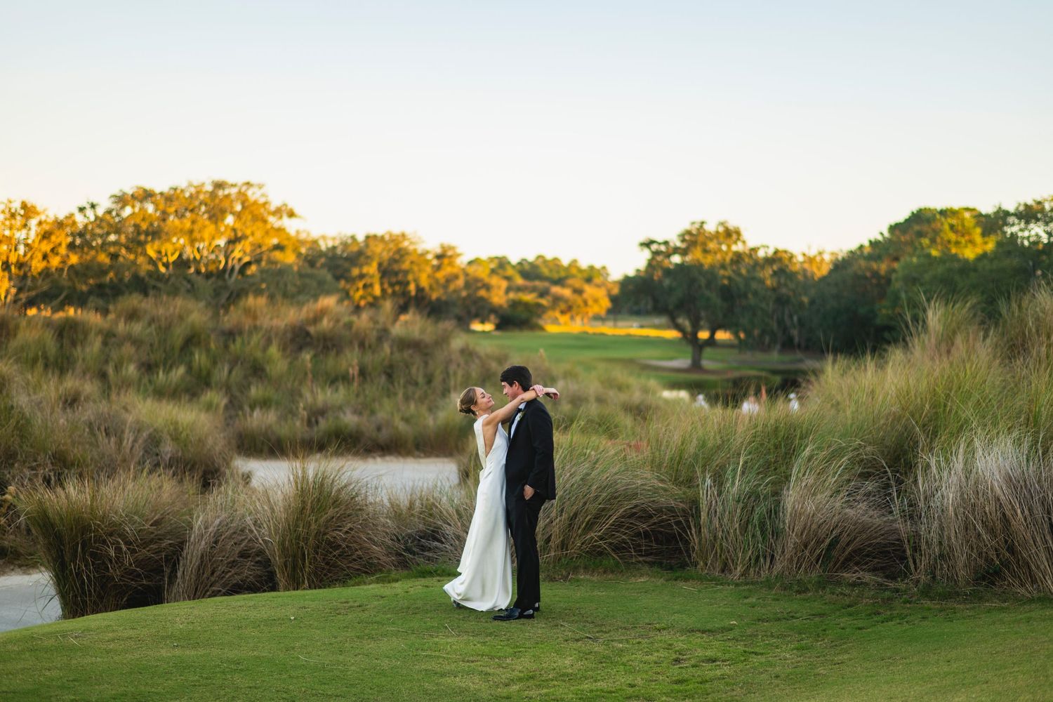 A couple in wedding attire embraces on a scenic golf course during golden hour sunset.