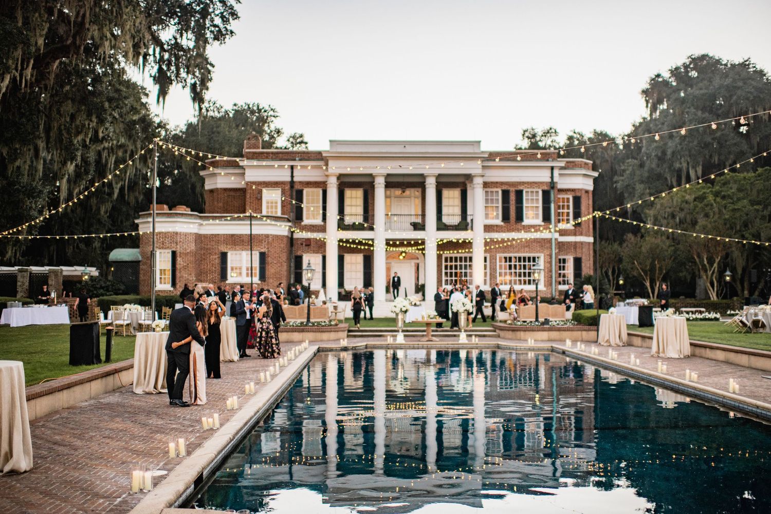 Elegant outdoor wedding venue with a grand pool and white columns illuminated at dusk with string lights overhead.
