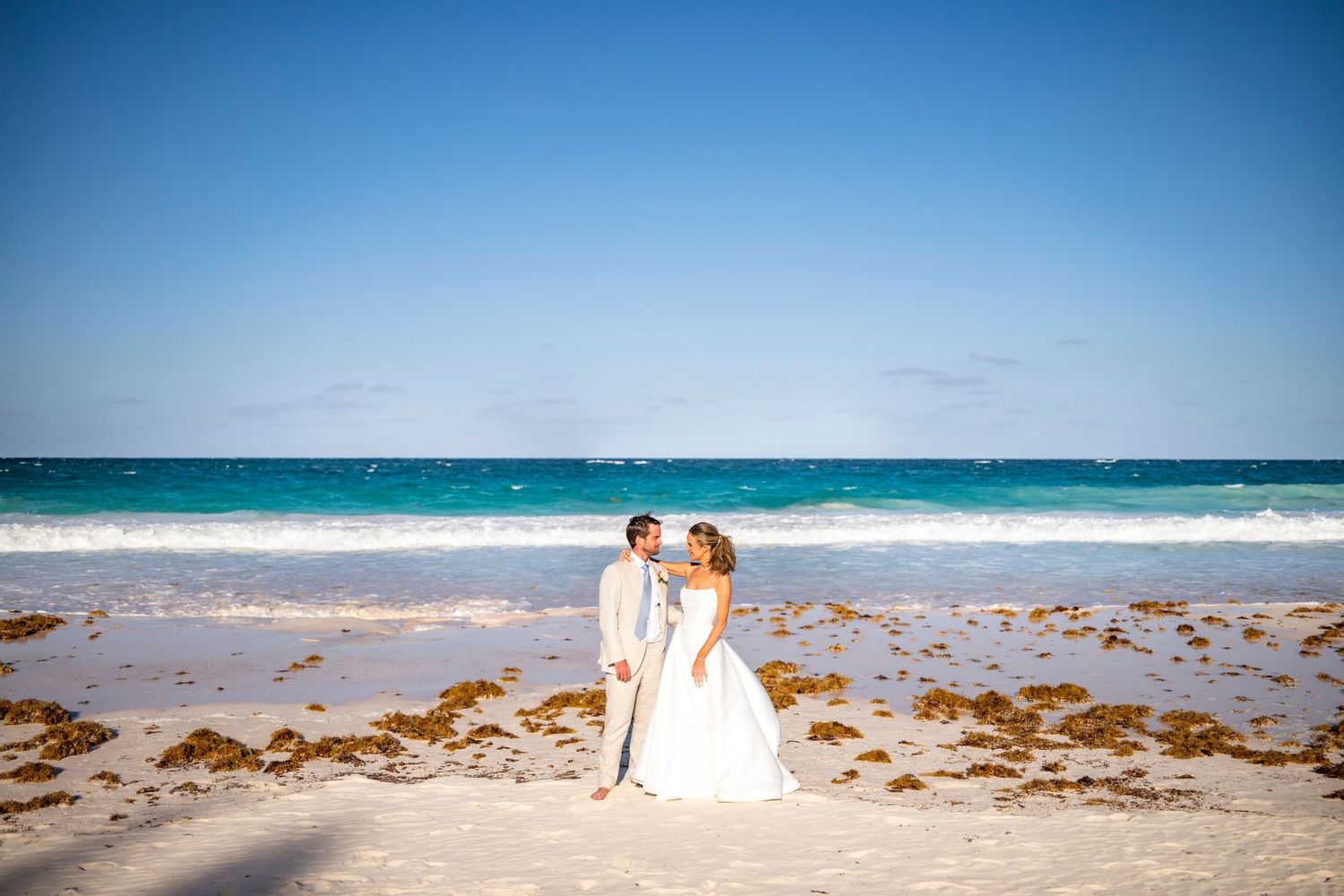 Wedding couple poses on sandy beach with turquoise ocean waves and blue sky in background.