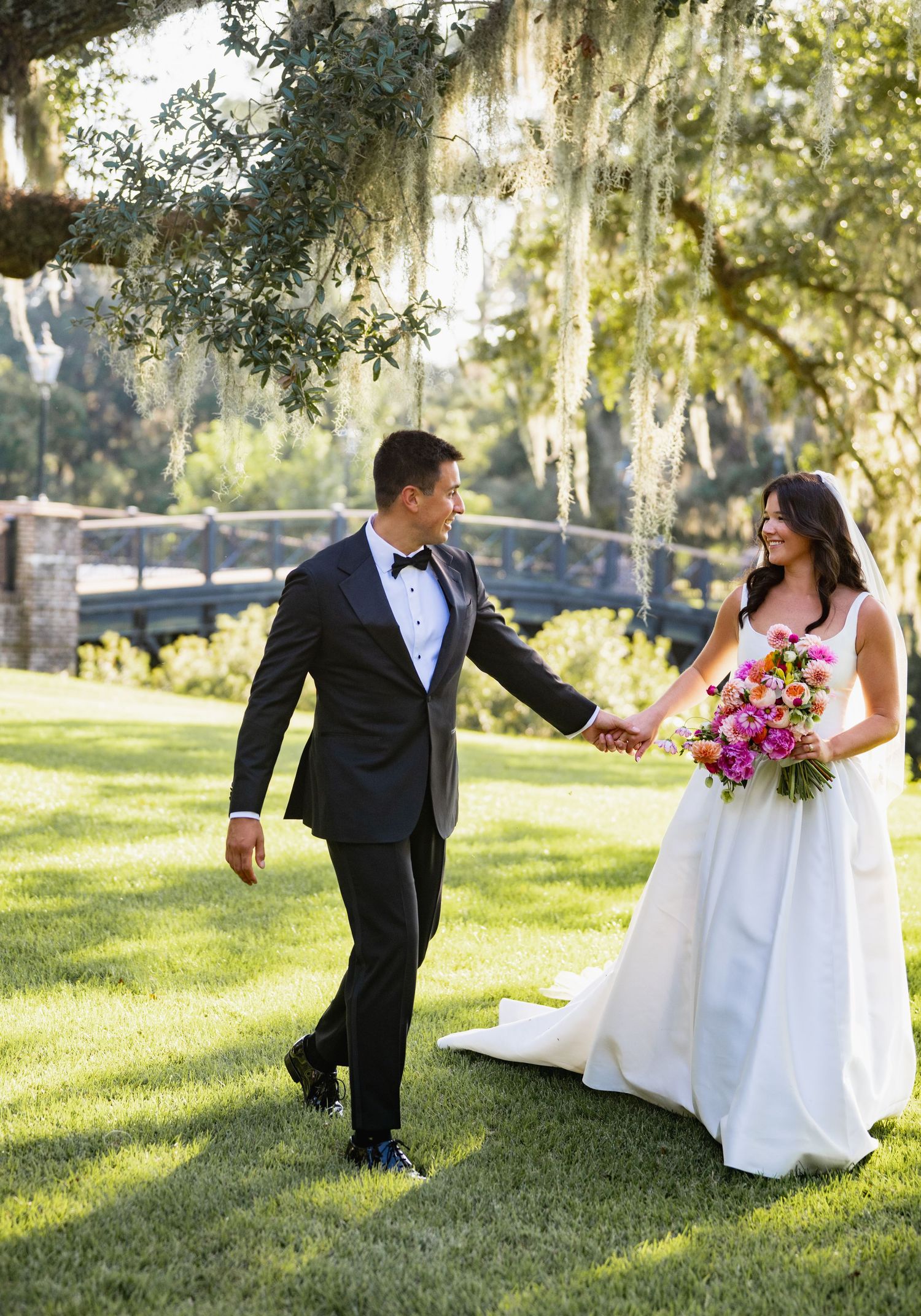 A couple in wedding attire playfully poses on a grassy lawn with Spanish moss hanging from trees in the background.
