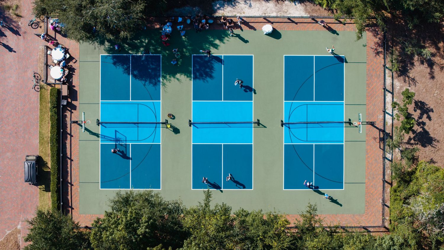 Aerial view of multiple blue tennis courts surrounded by trees and walkways at a sports facility.