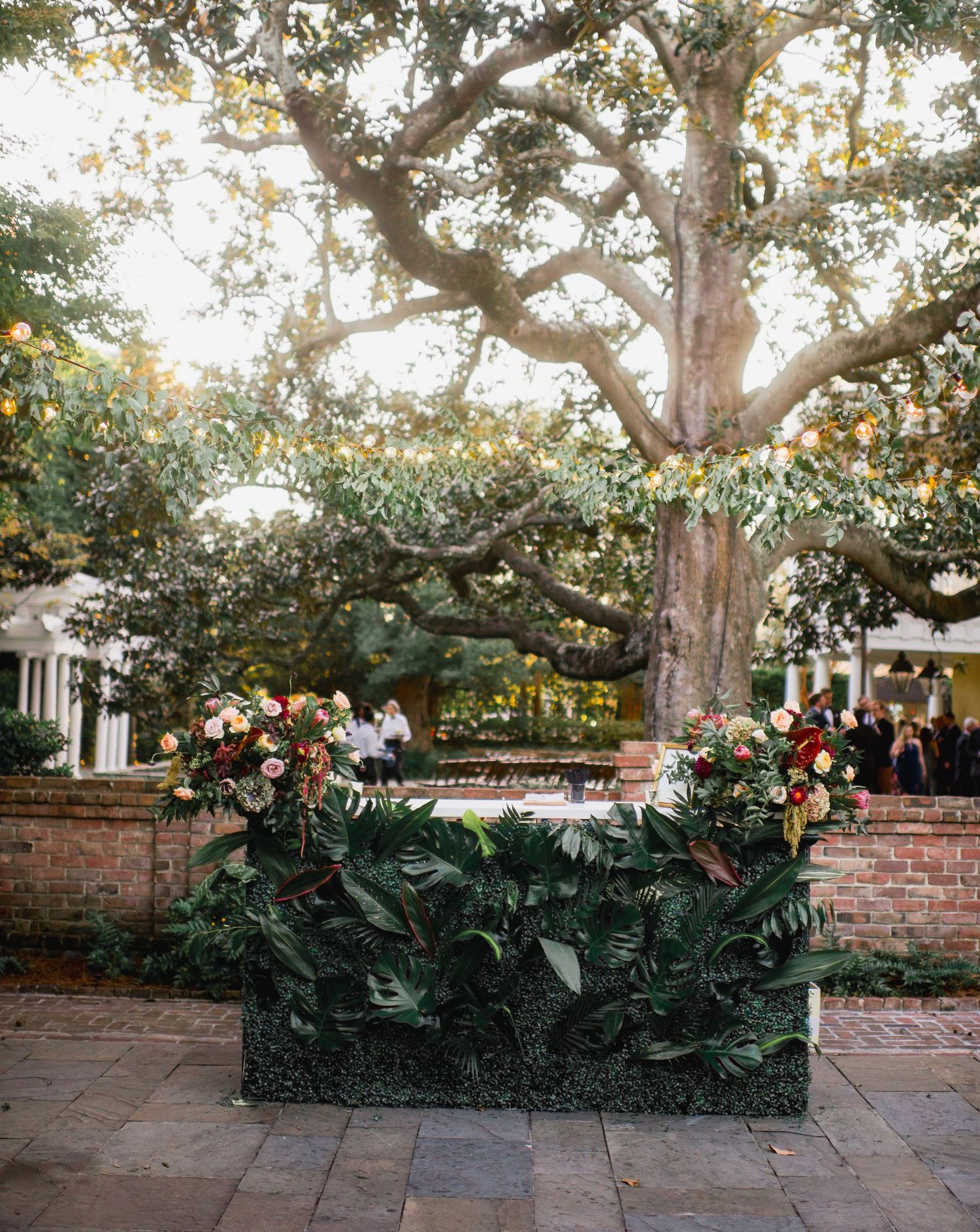 Tropical foliage and floral arrangements adorn a brick wall and patio area in an outdoor garden setting.