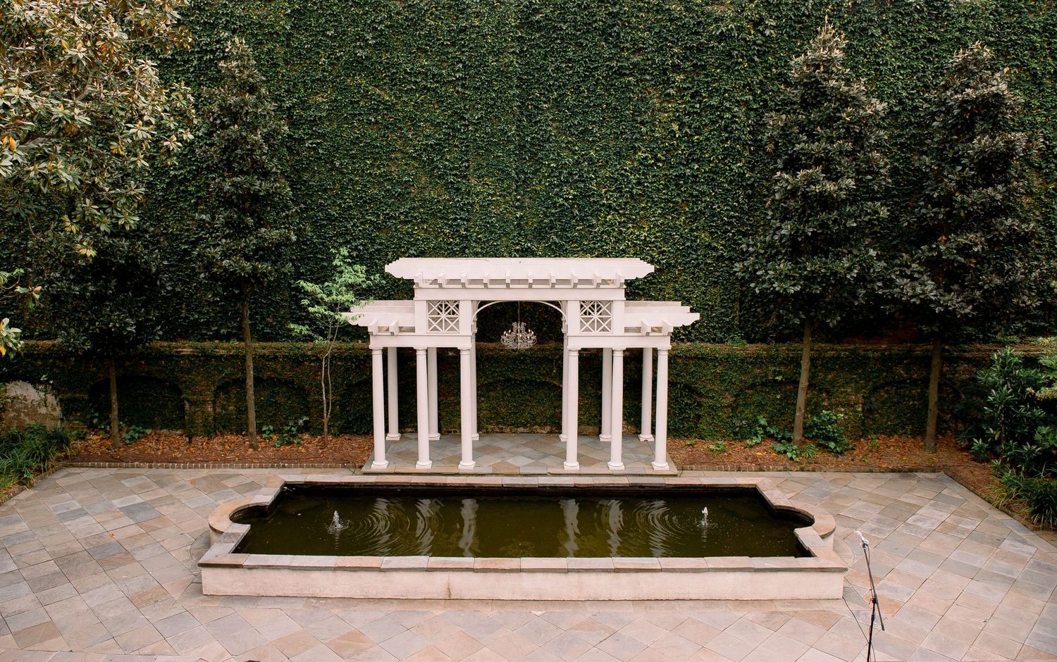 Elegant white marble pergola with columns stands over a reflecting pool in a lush garden setting.