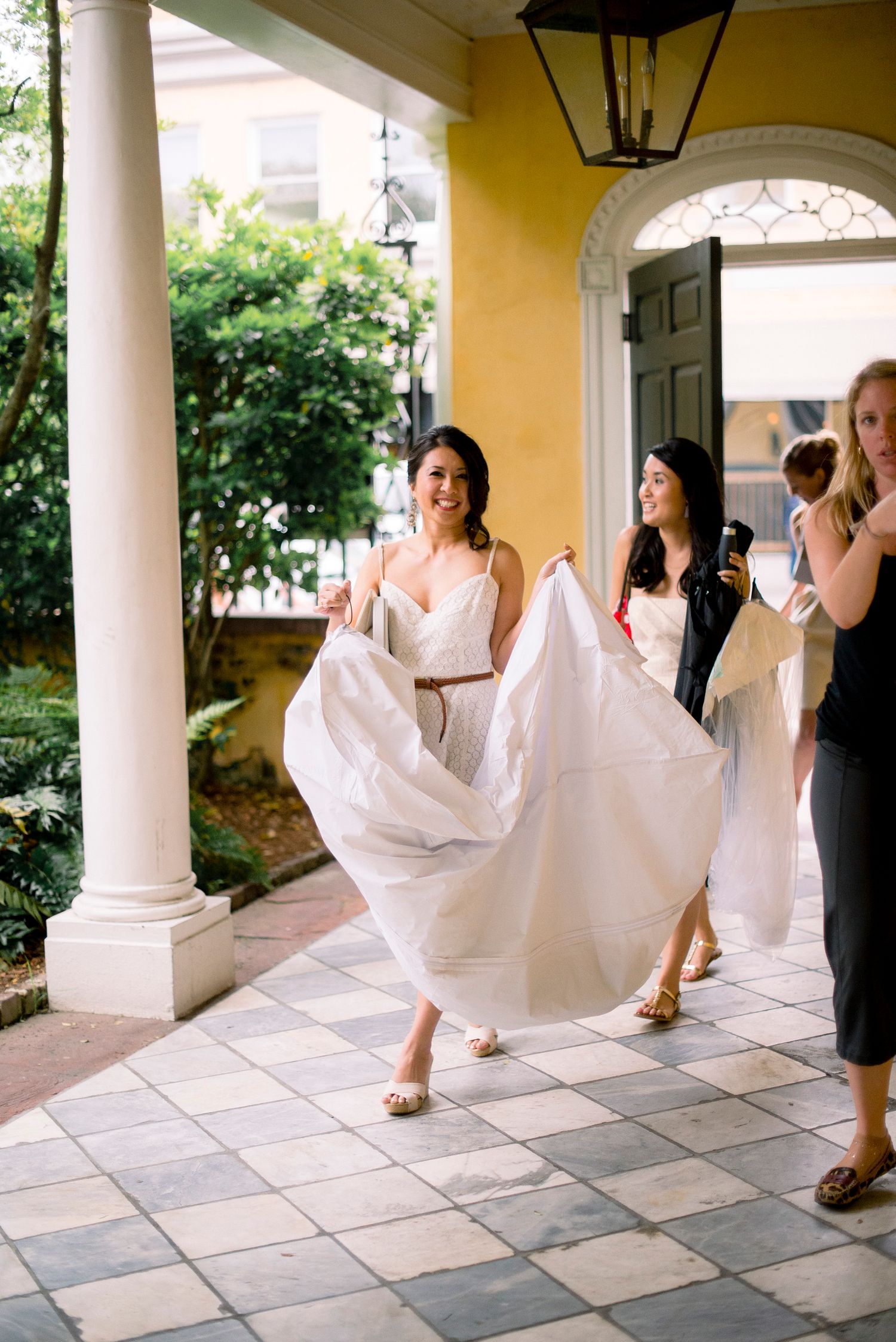 A bridal party in white dresses walking on a tiled porch with elegant columns and yellow walls in the background.