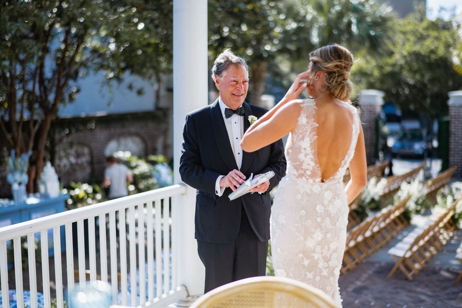 A wedding couple shares an intimate moment on a white porch surrounded by sunlit trees and gardens.