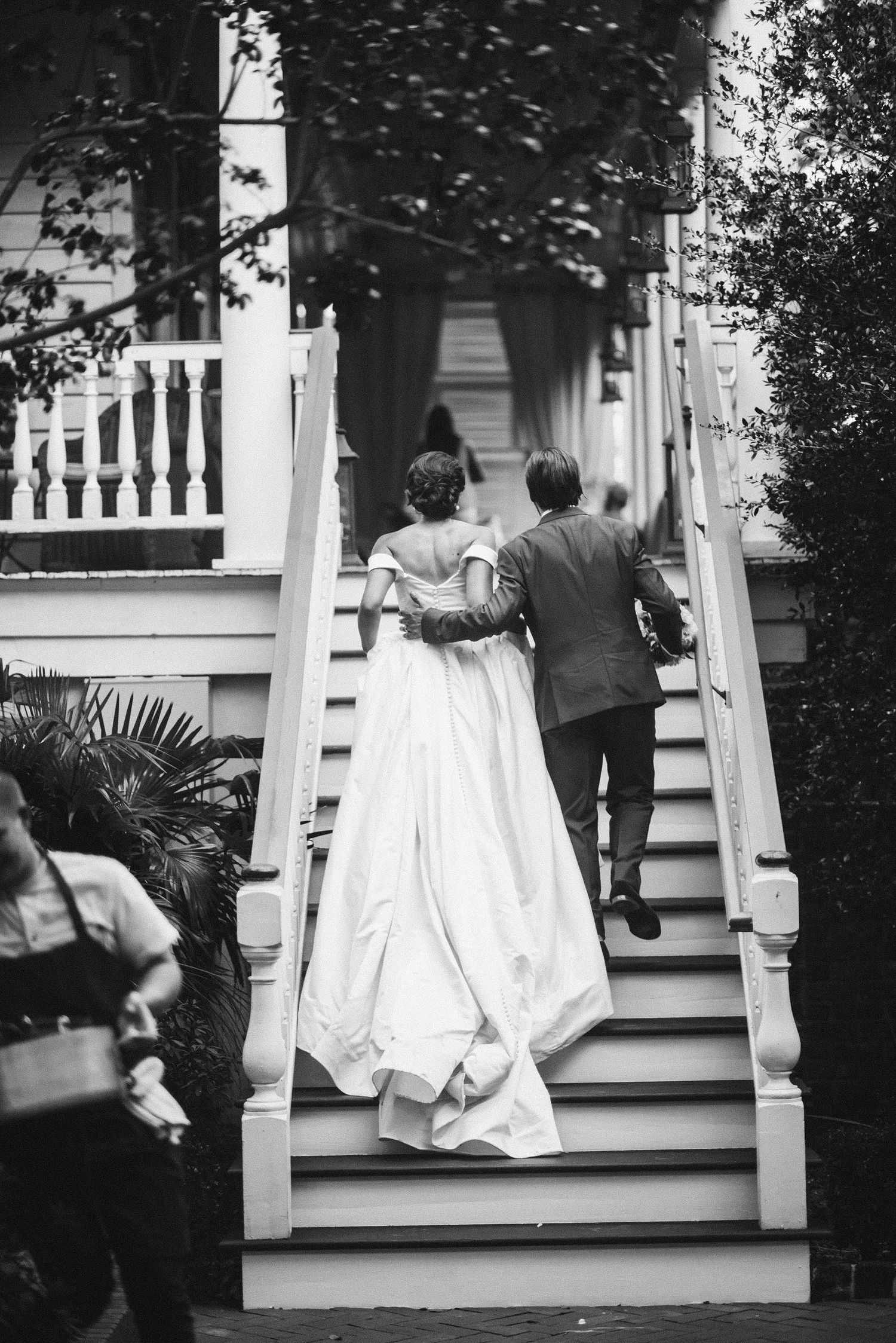 A couple ascends white wooden stairs in formal wedding attire, captured in black and white photography.