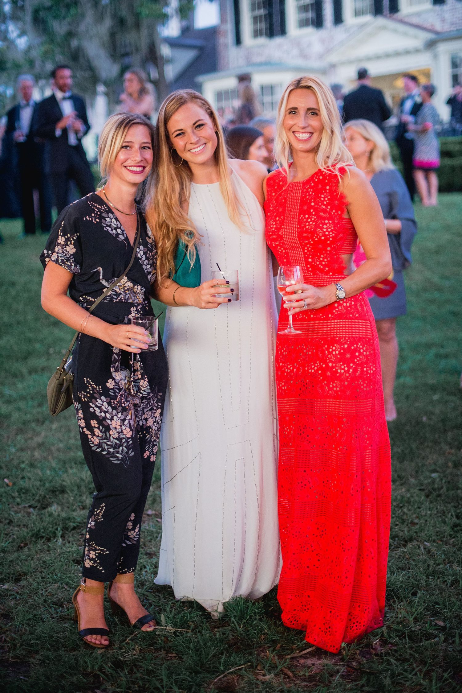 Three women in evening attire pose together at an outdoor event, wearing black, white and red dresses.
