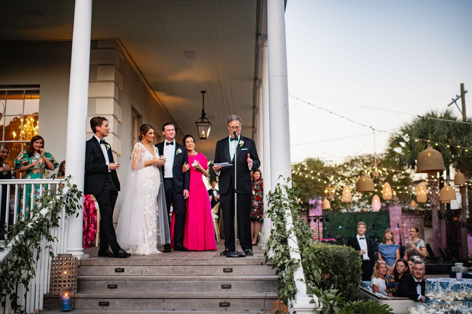 A wedding party gathers on a decorated porch at dusk while guests watch from the lawn below.