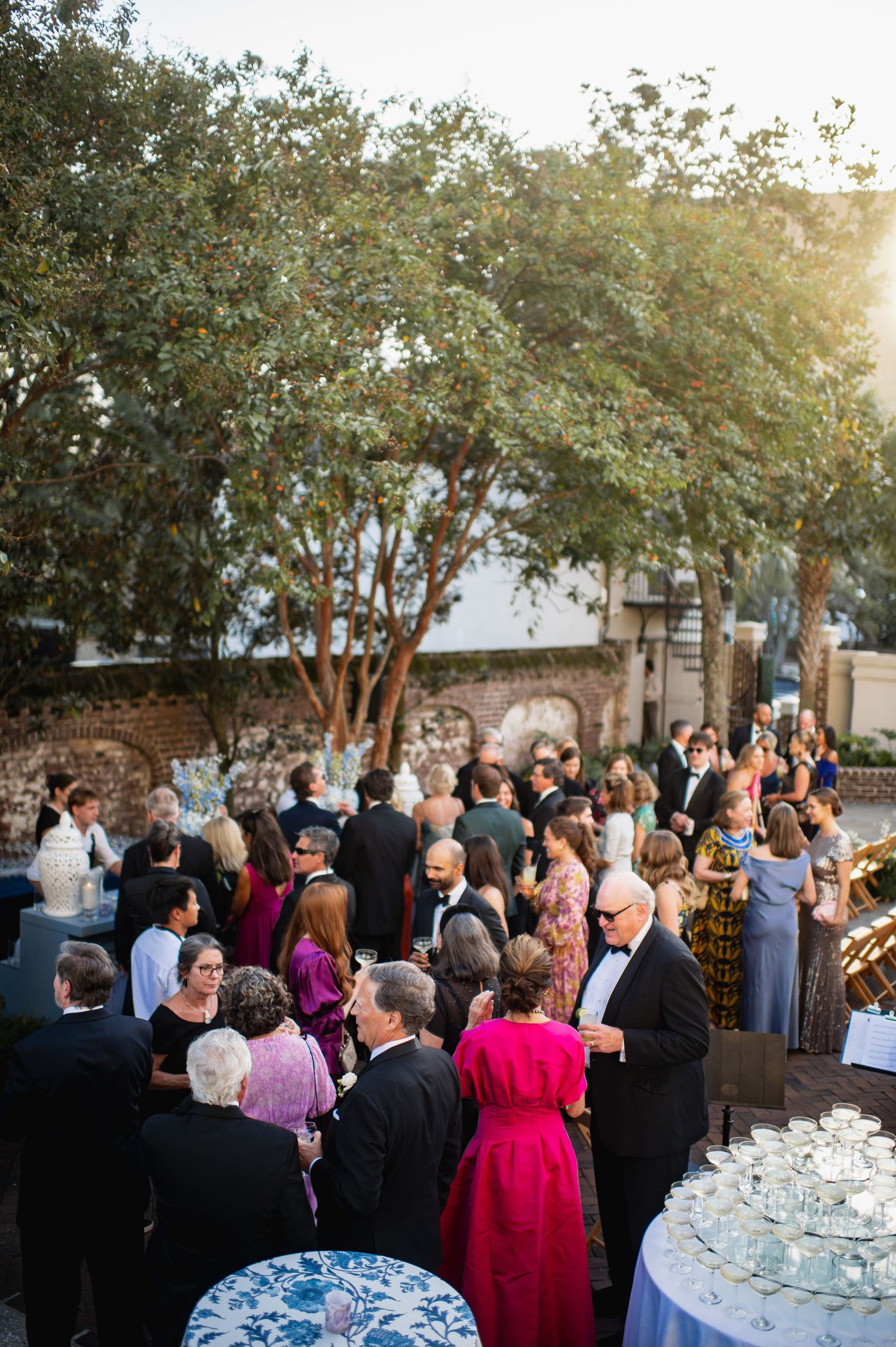 Guests gather at an outdoor cocktail reception under trees at sunset with wine glasses and tables.