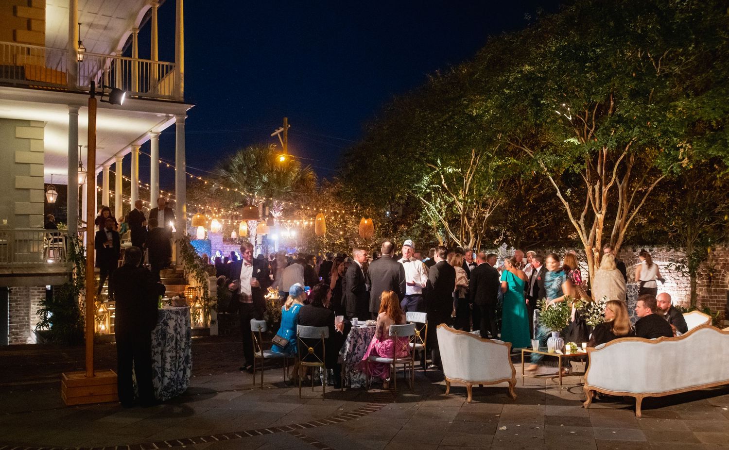 A panoramic view of a nighttime street festival with glowing lights, trees, and people gathering along the sidewalks.