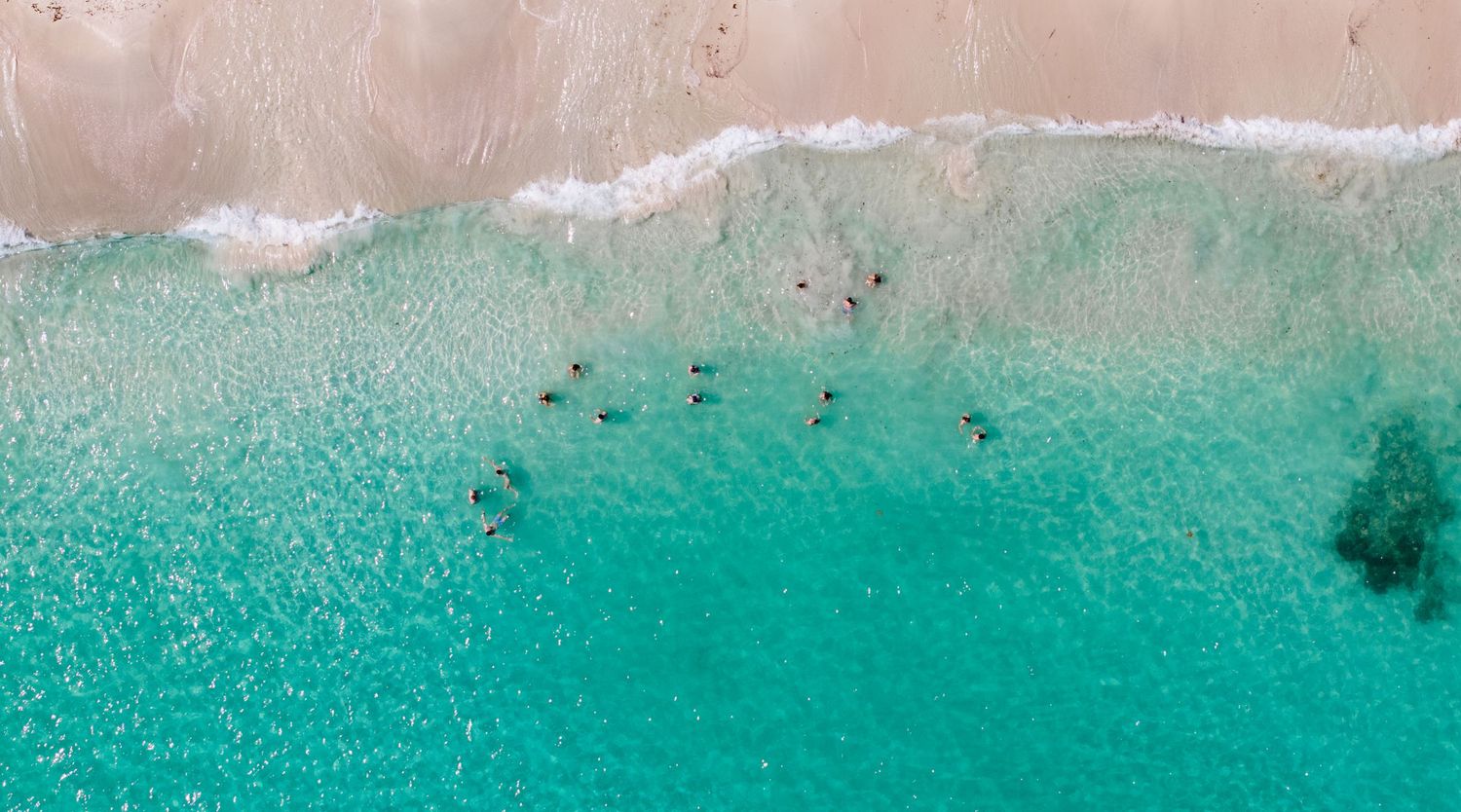 Aerial view of turquoise ocean waves meeting sandy beach with dark spots visible in crystal clear waters.